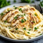 A close-up view of creamy fettuccine Alfredo pasta topped with perfectly grilled chicken slices, garnished with fresh parsley and grated parmesan cheese, served in an elegant white bowl with a fork on the side.