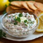 A glass bowl of smoked fish dip garnished with parsley and a lemon slice, surrounded by toasted bread slices and a lemon wedge in a cozy kitchen setting.